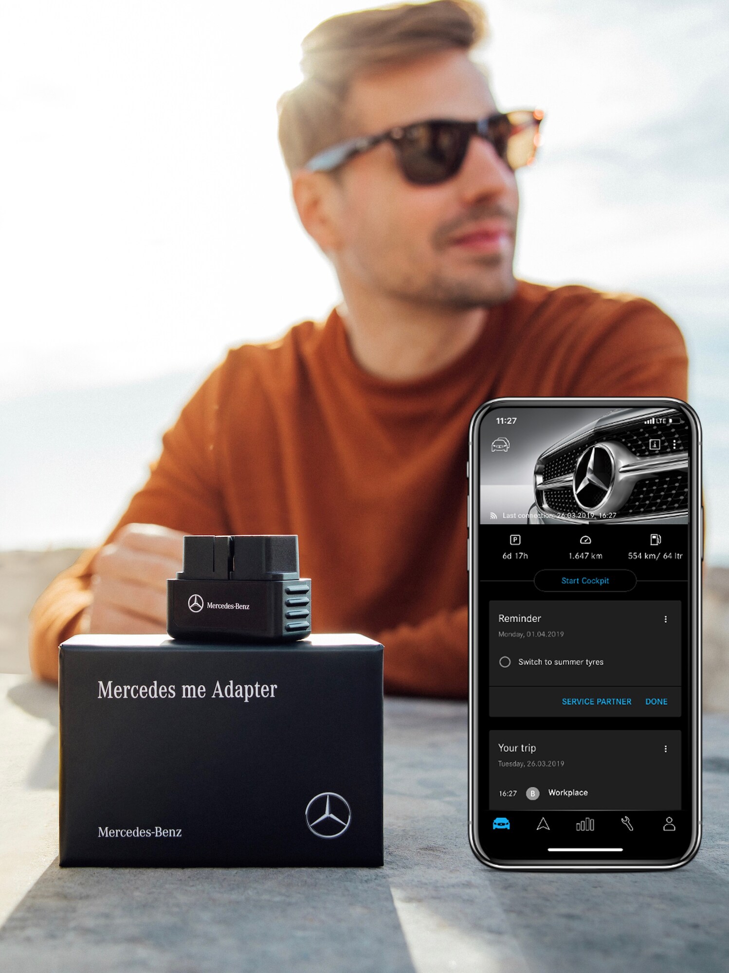 Een man met een zonnebril zit aan een tafel, waarbij op de voorgrond een Mercedes-Benz Adapter en smartphone met geopende Mercedes-Benz Adapter-app liggen.