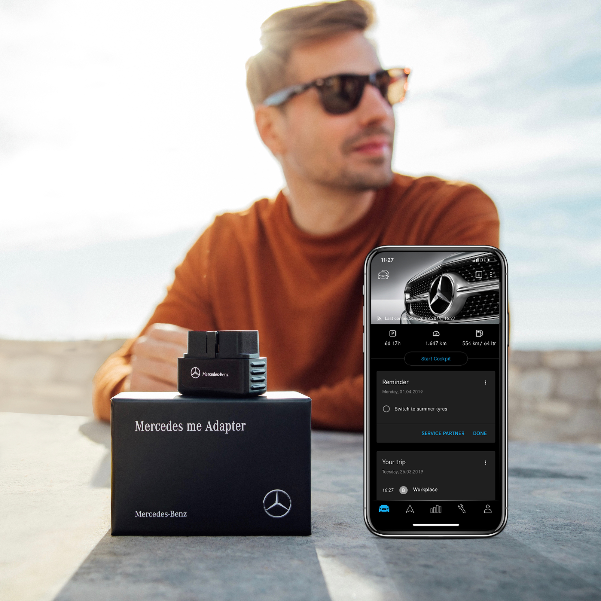 Ongevaldetectie met de Mercedes-Benz Adapter. | Mercedes-Benz Een man met een zonnebril zit aan een tafel, waarbij op de voorgrond een Mercedes-Benz Adapter en smartphone met geopende Mercedes-Benz Adapter-app liggen.