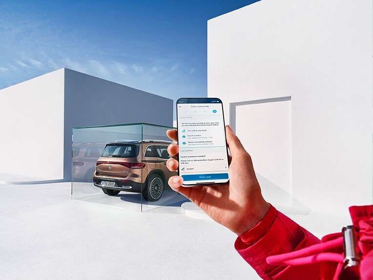 Iemand met een mobiele telefoon in de hand die de Mercedes App gebruikt. Op de achtergrond wordt een EQB geparkeerd.