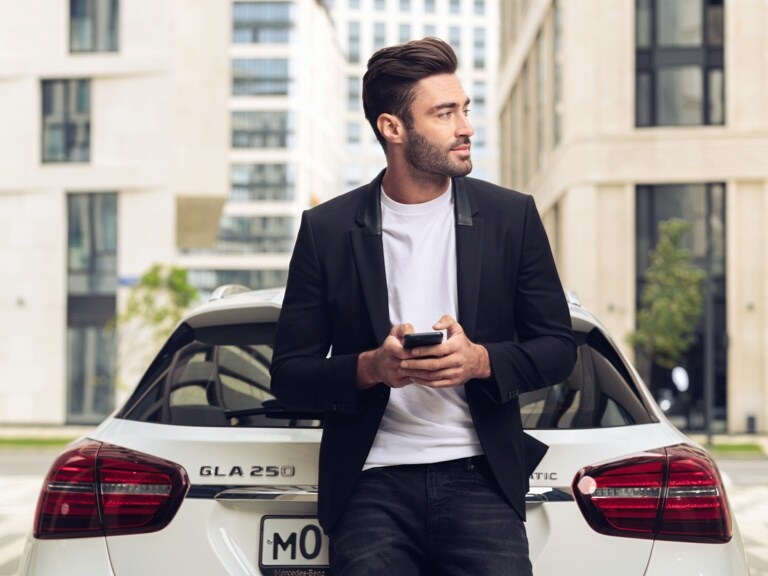 Een man leunt tegen zijn Mercedes-Benz GLA en laat zich via de Mercedes-Benz App op zijn smartphone en de digitale extra's voor navigatie informeren wat de weersverwachting op de bestemming is.