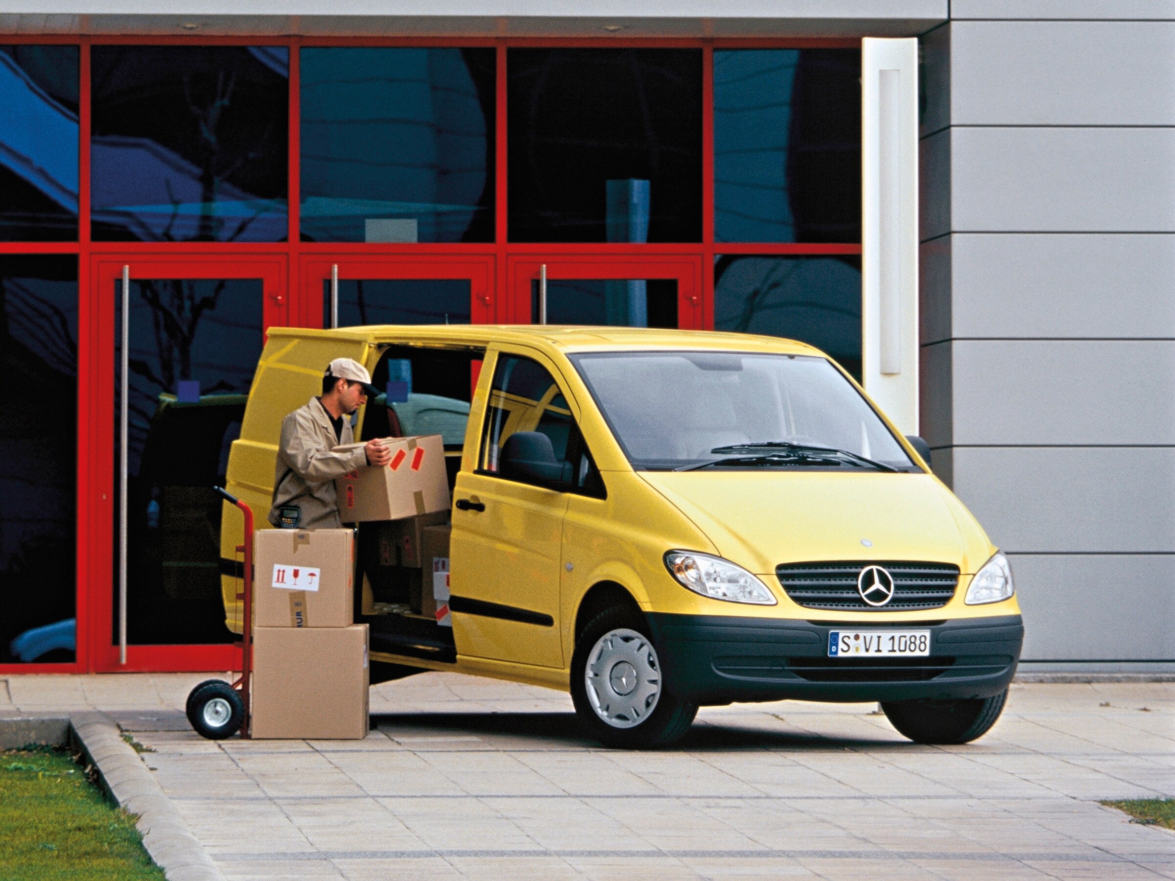 Een gele Mercedes-Benz Vito staat geparkeerd voor het gebouw, terwijl een medewerker pakketten uitlaadt