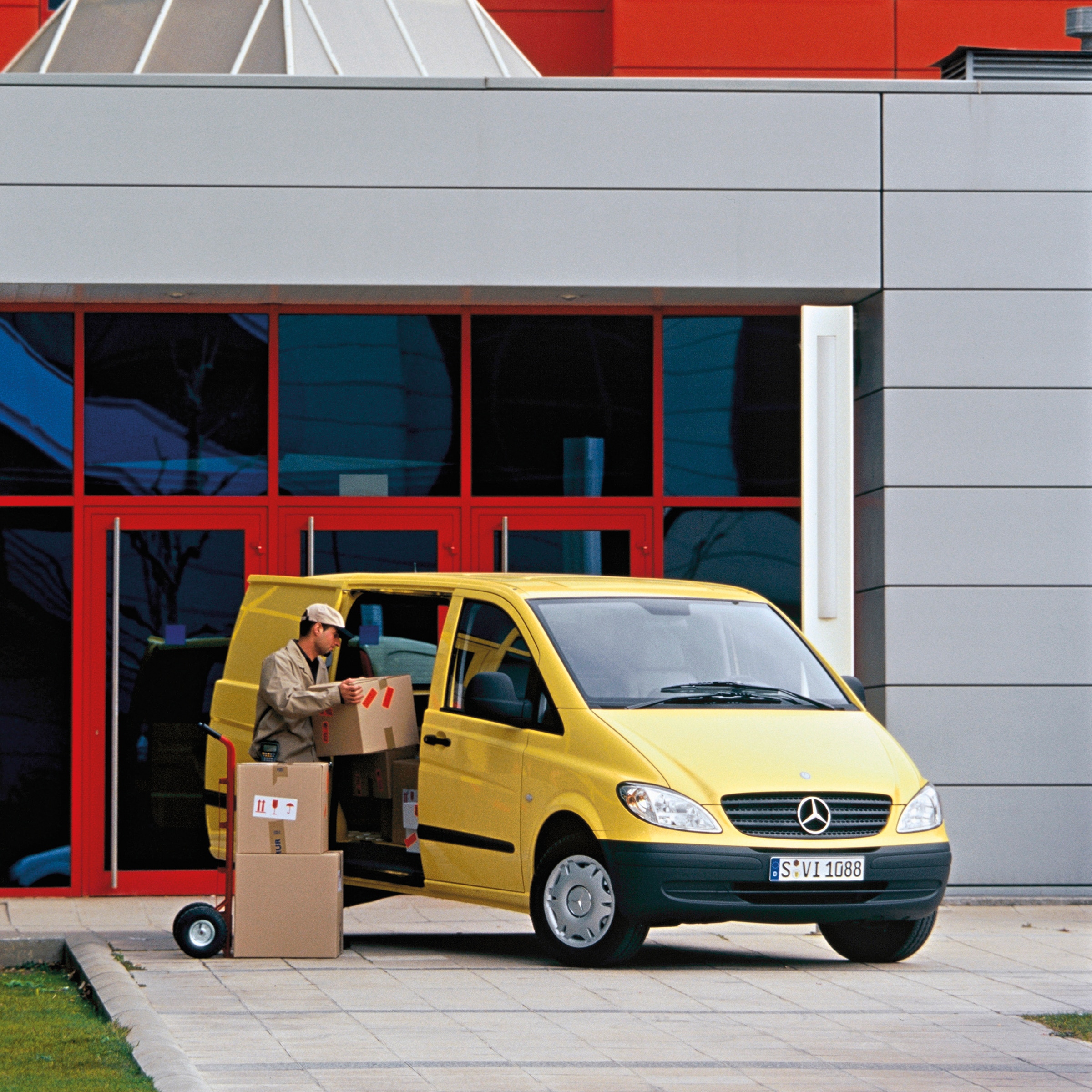 De tweede generatie Mercedes-Benz Vito’s | Geschiedenis van bedrijfswagens | Mercedes-Benz Een gele Mercedes-Benz Vito staat geparkeerd voor het gebouw, terwijl een medewerker pakketten uitlaadt