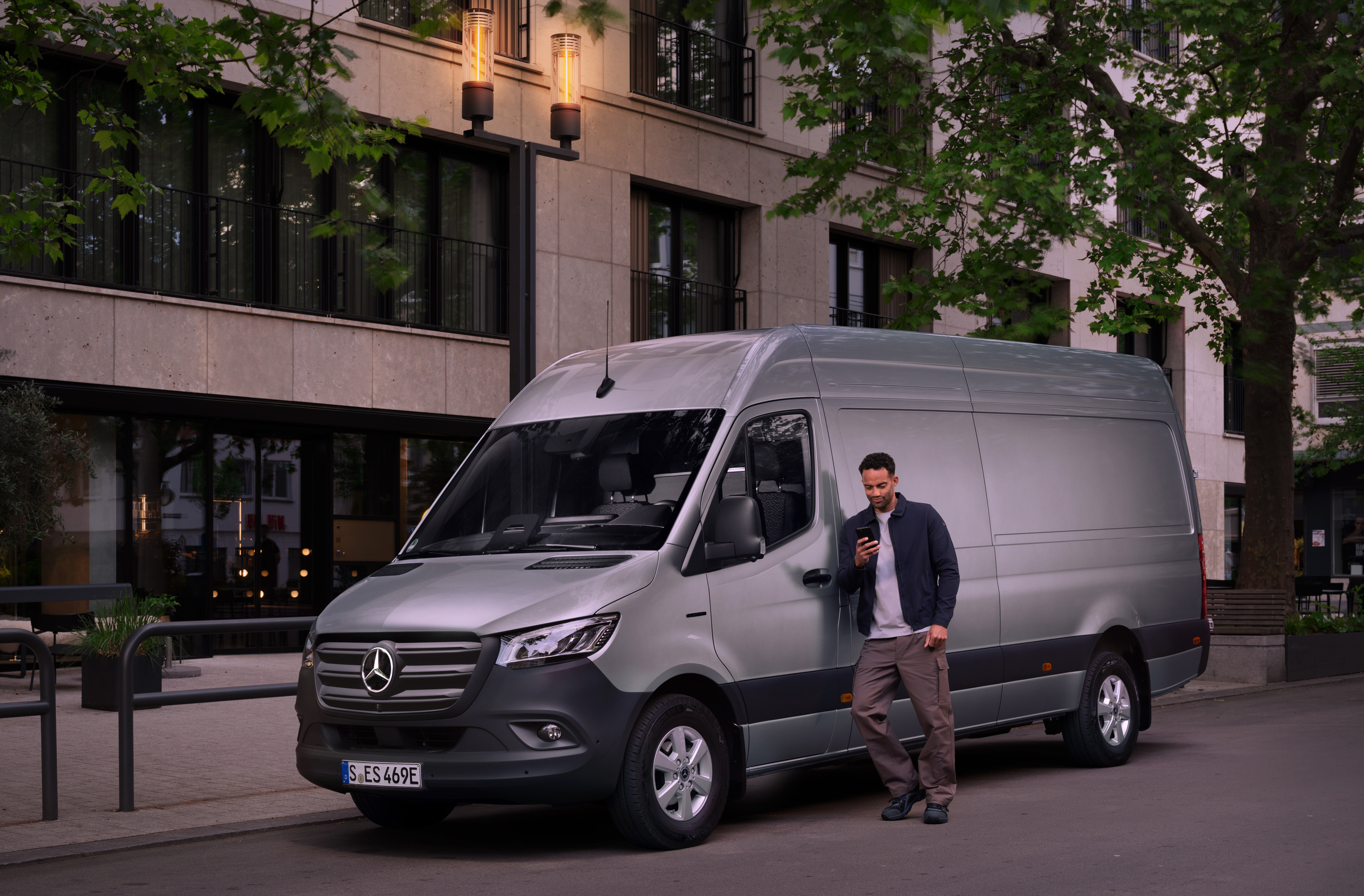 Mercedes-Benz Bedrijfswagen | Vans Care | Customer Assistance Center Man leunt tegen een zilveren Mercedes-Benz Sprinter Gesloten Bestelwagen en gebruikt zijn smartphone – snelle hulp van Mercedes-Benz Klantenzorg.