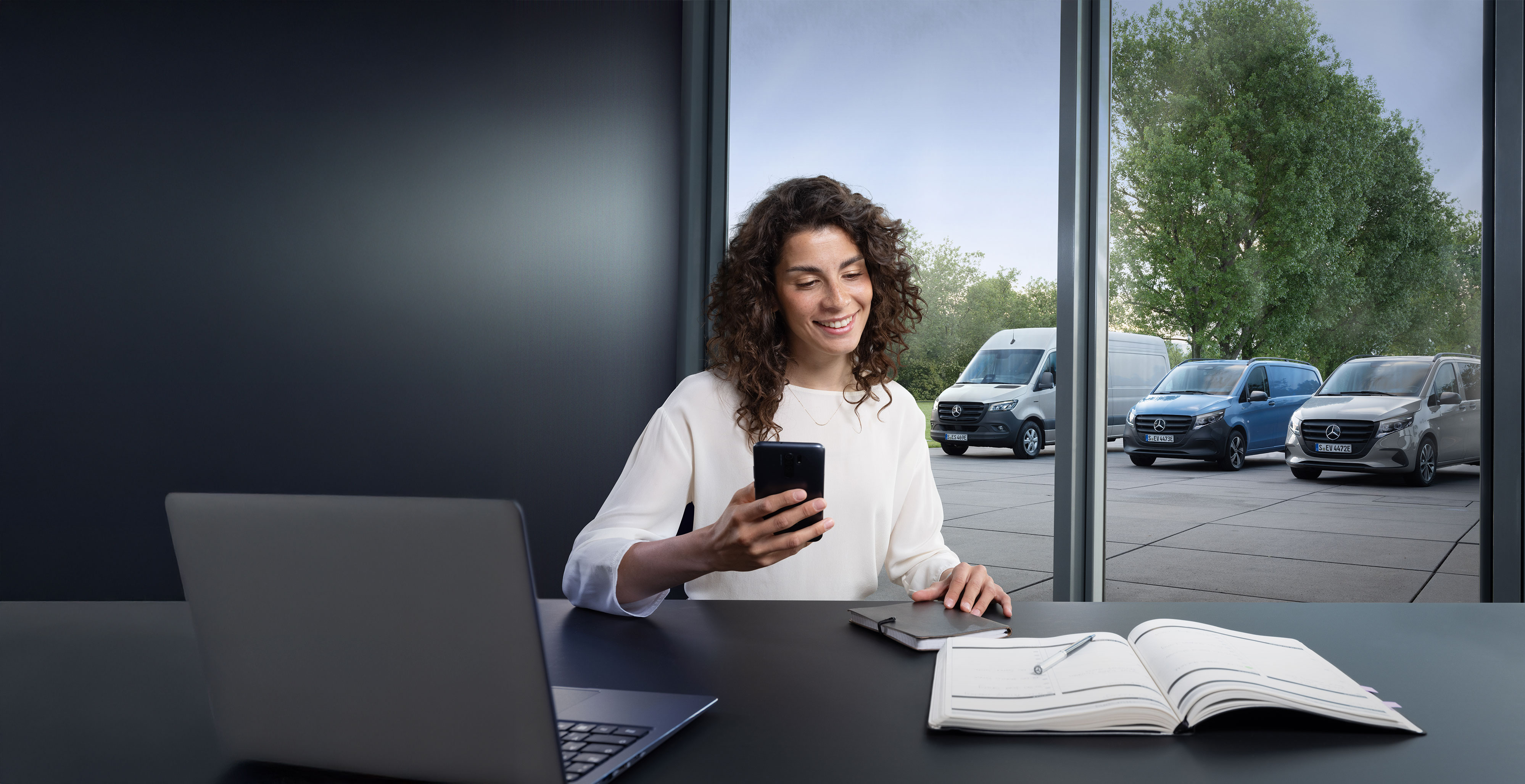 Mercedes-Benz Bedrijfswagen | Vans Care Vrouw met smartphone aan een bureau, met meerdere Mercedes-Benz Bedrijfswagens op de achtergrond – deskundige ondersteuning in Business Care.