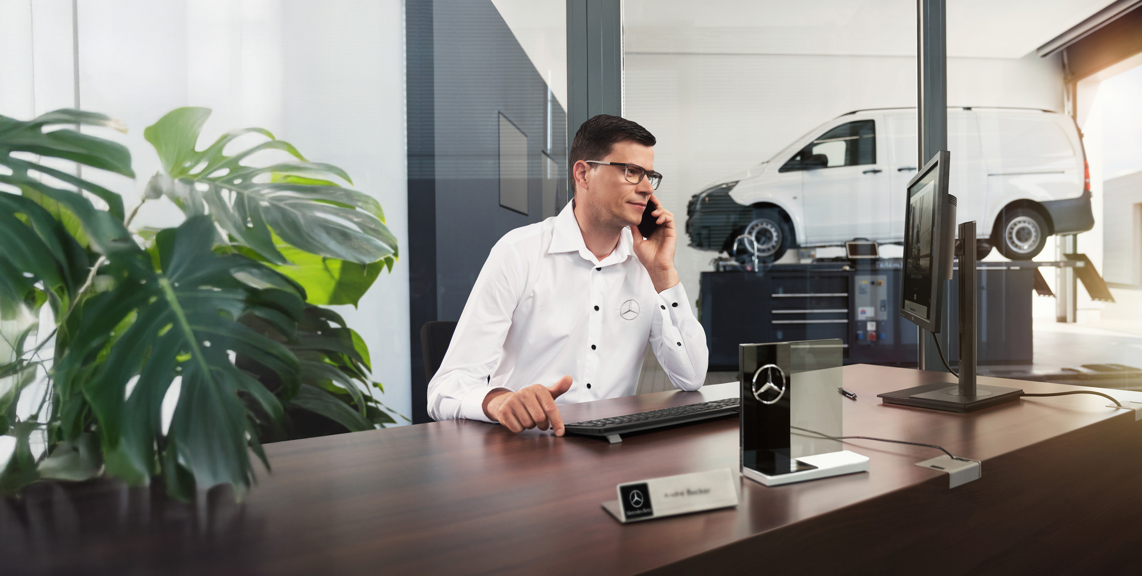 Mercedes-Benz | Vans Care Mercedes-Benz medewerker telefoneert aan een bureau met een Mercedes-Benz Vito bestelwagen in de werkplaats.