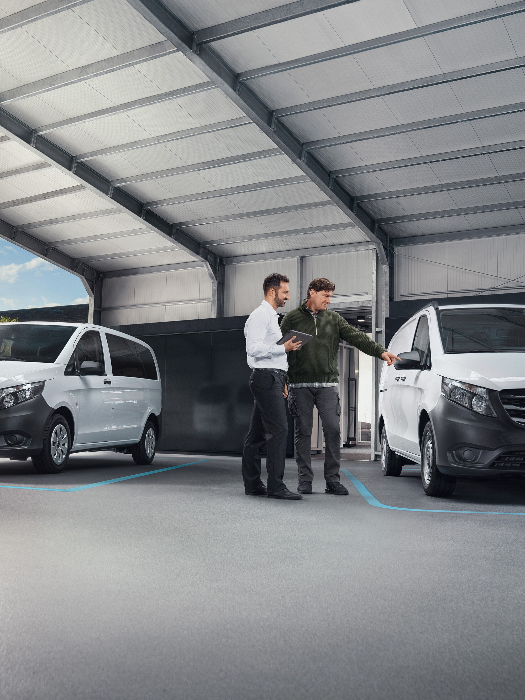 Twee personen in een werkplaats in gesprek, staand voor twee witte Mercedes-Benz bestelwagens.