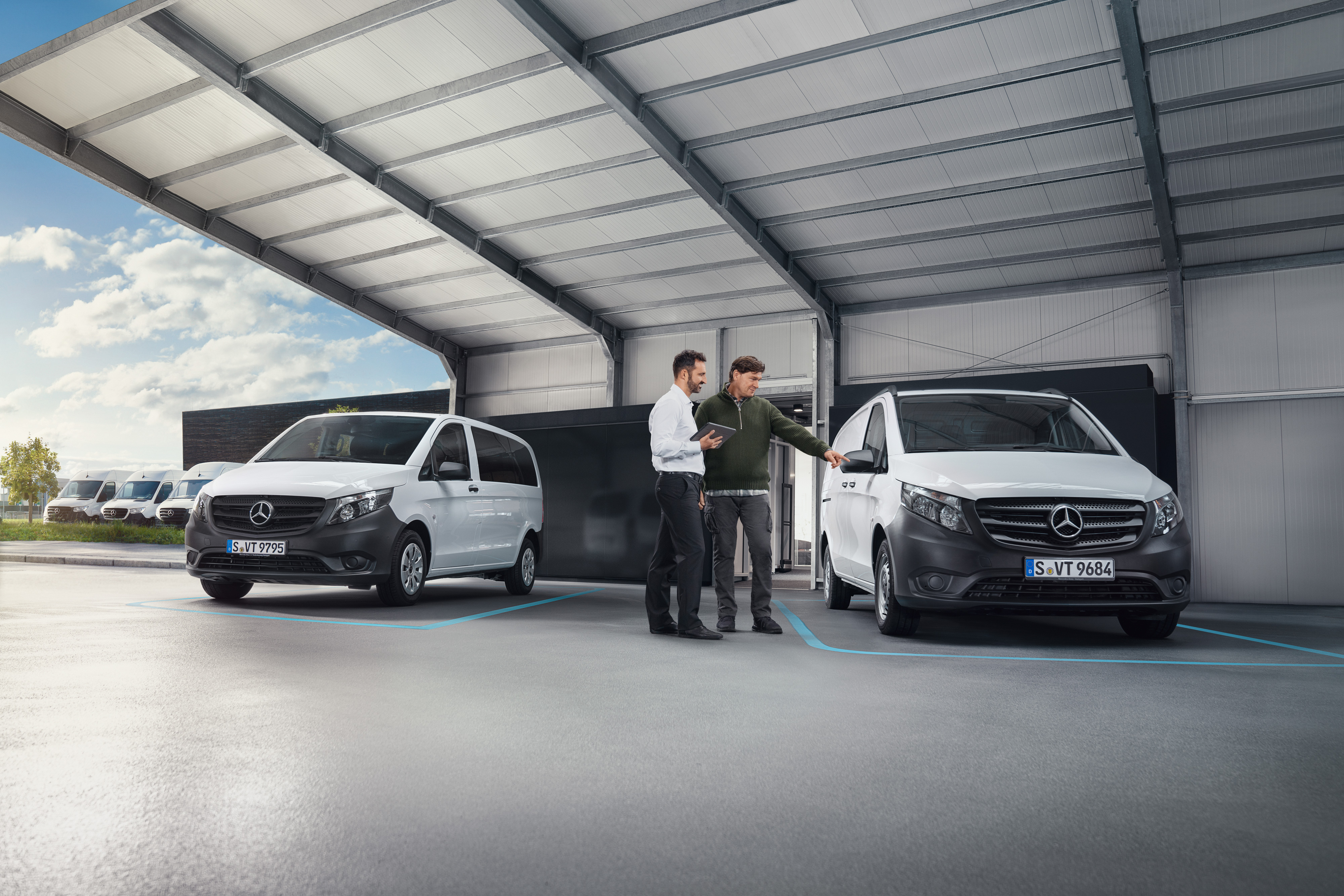 Mercedes-Benz | Comfort | Digitale klantenreceptie Twee personen in een werkplaats in gesprek, staand voor twee witte Mercedes-Benz bestelwagens.