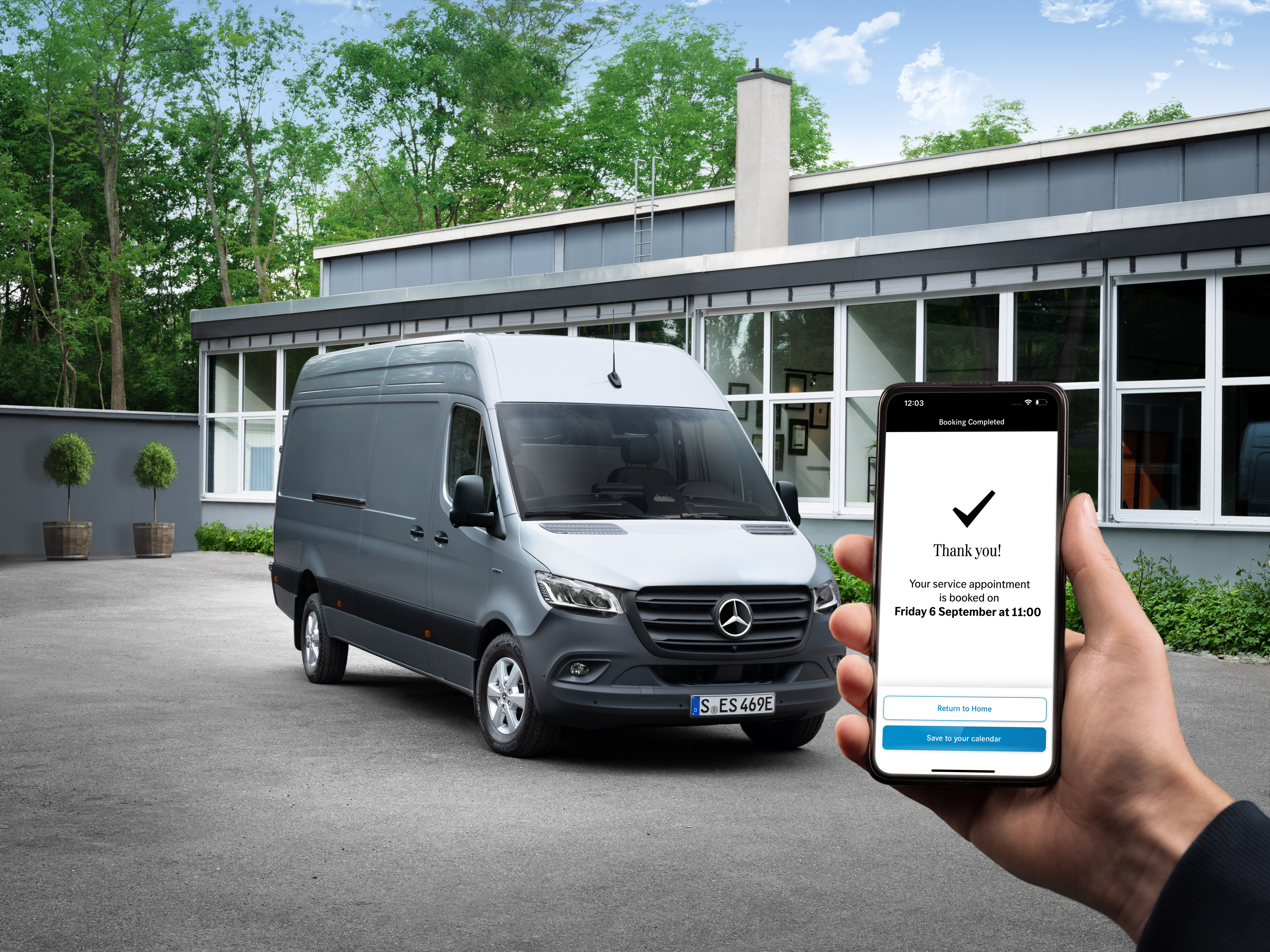 Mercedes-Benz | Comfort | Service Partner Een grijze Mercedes-Benz Sprinter bestelwagen voor een kantoor, met op de voorgrond een persoon die een smartphone vasthoudt.
