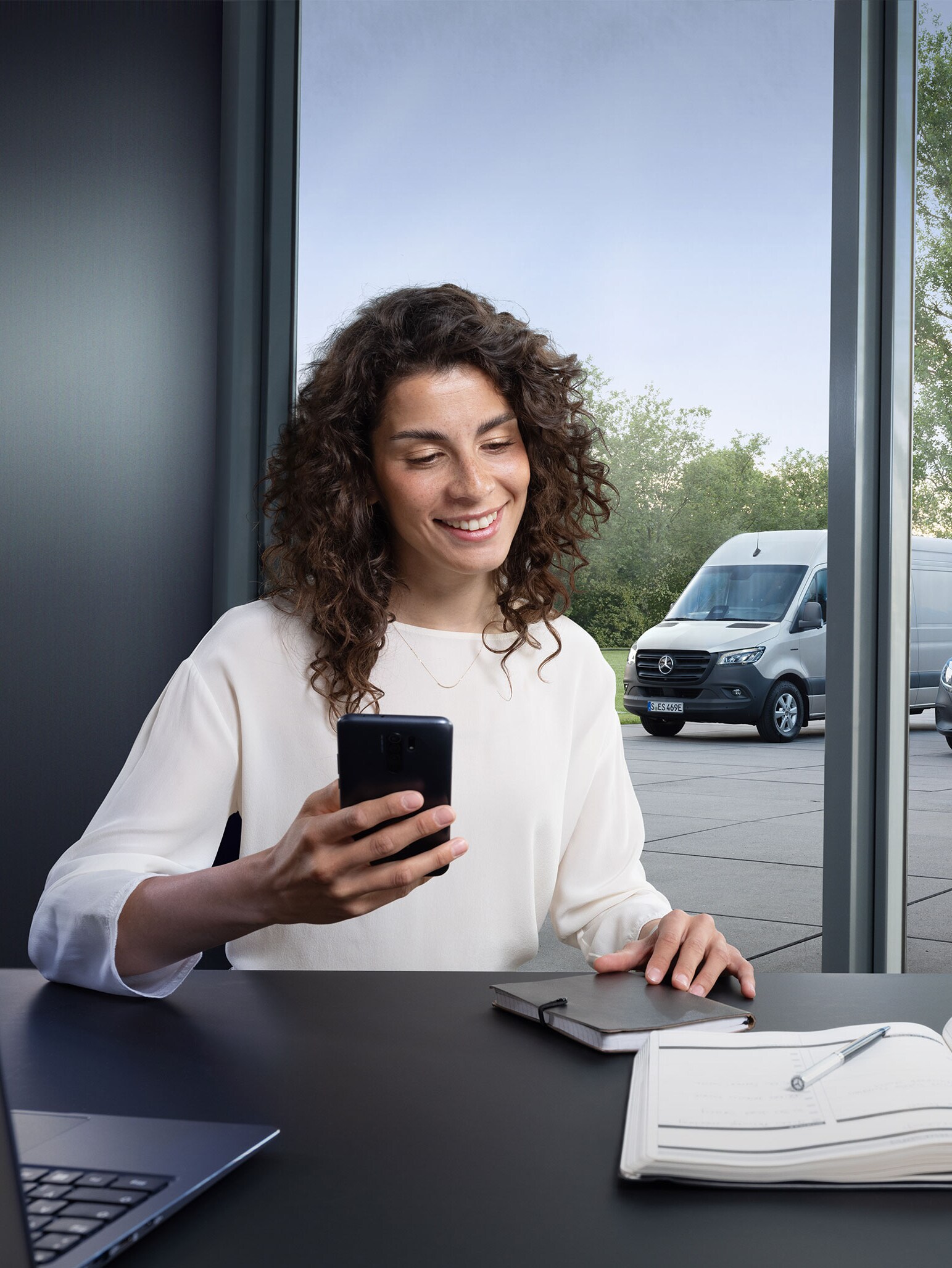 Vrouw boekt een service via smartphone met een reeks Mercedes-Benz bestelwagens op de achtergrond.