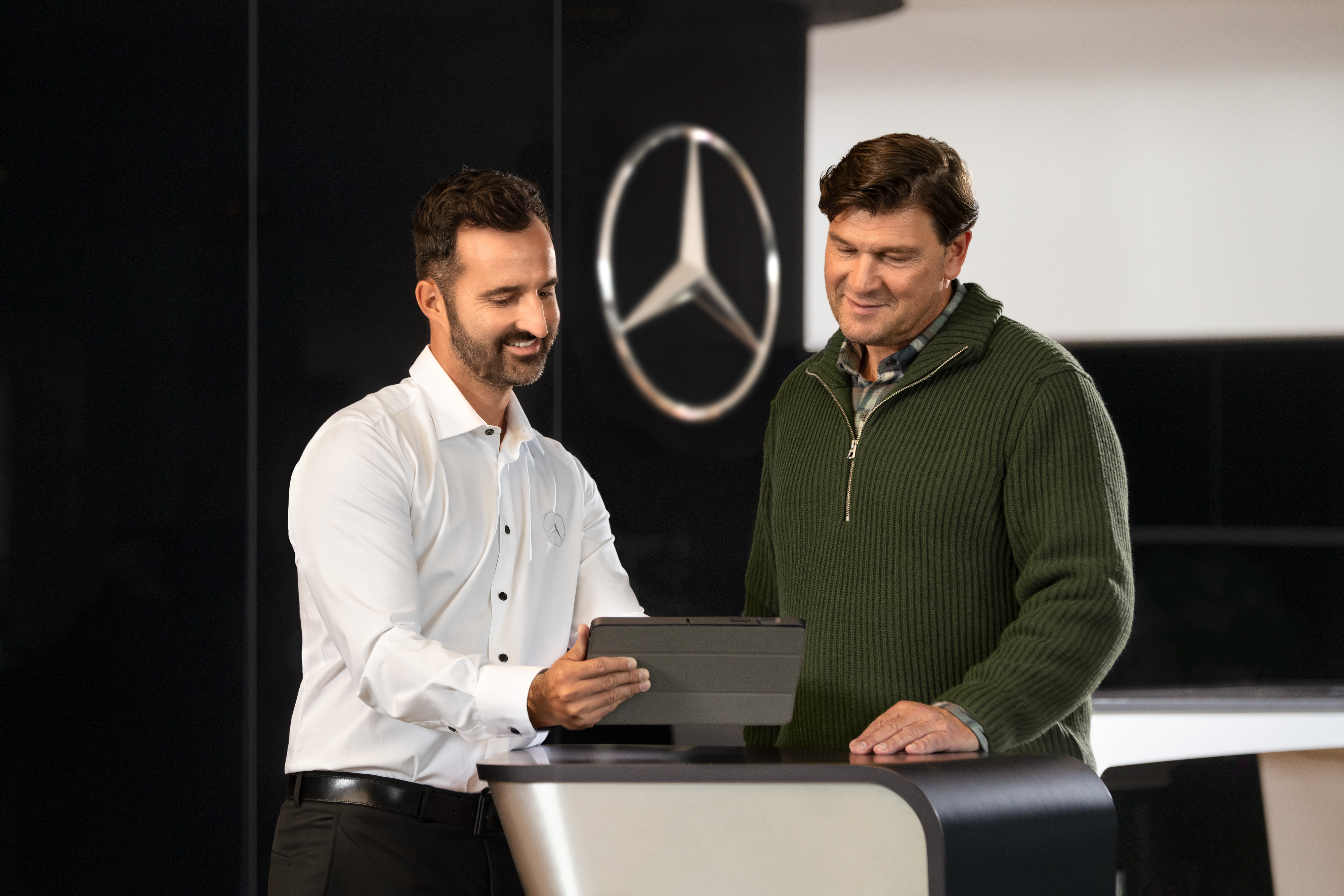 Mercedes-Benz | Servicesoverzicht Twee mannen praten aan een balie terwijl ze op een tablet kijken, voor de ster van Mercedes-Benz.