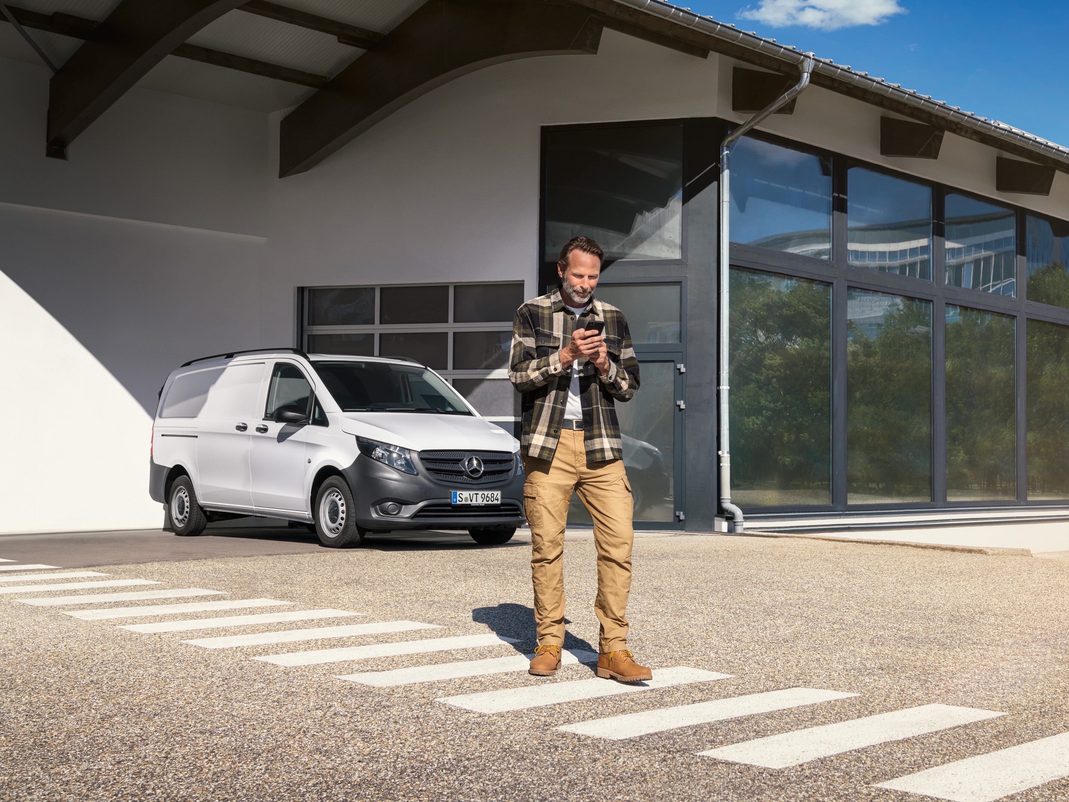 Een man die voor een witte Mercedes-Benz Vito Gesloten Bestelwagen staat en zijn smartphone gebruikt.