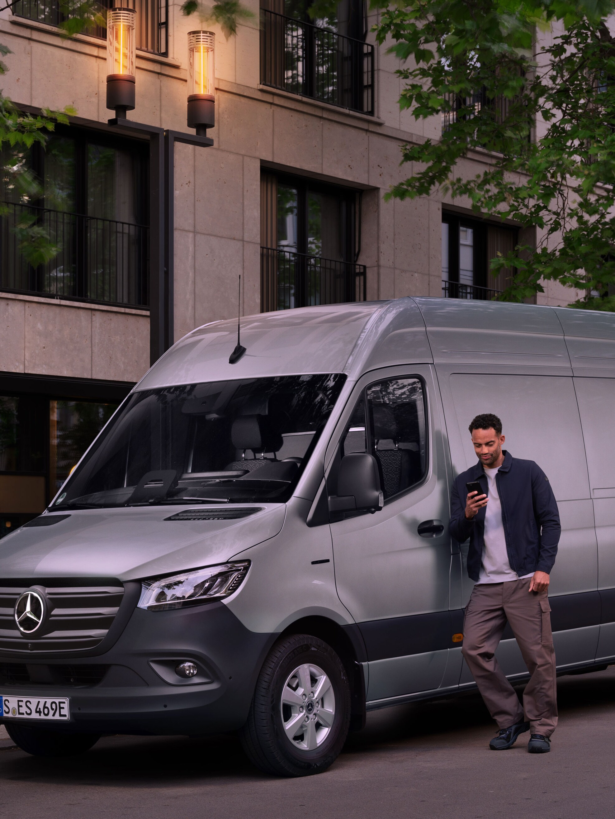 Man leunt tegen een zilveren Mercedes-Benz Sprinter en vult een schaderapport in op zijn mobiele telefoon.