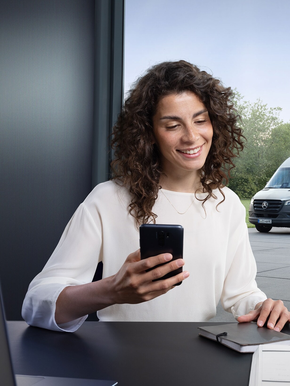 Vrouw die met een smartphone het vlootbeheer van meerdere Mercedes-Benz bedrijfswagens bedient bij het kantoorraam.