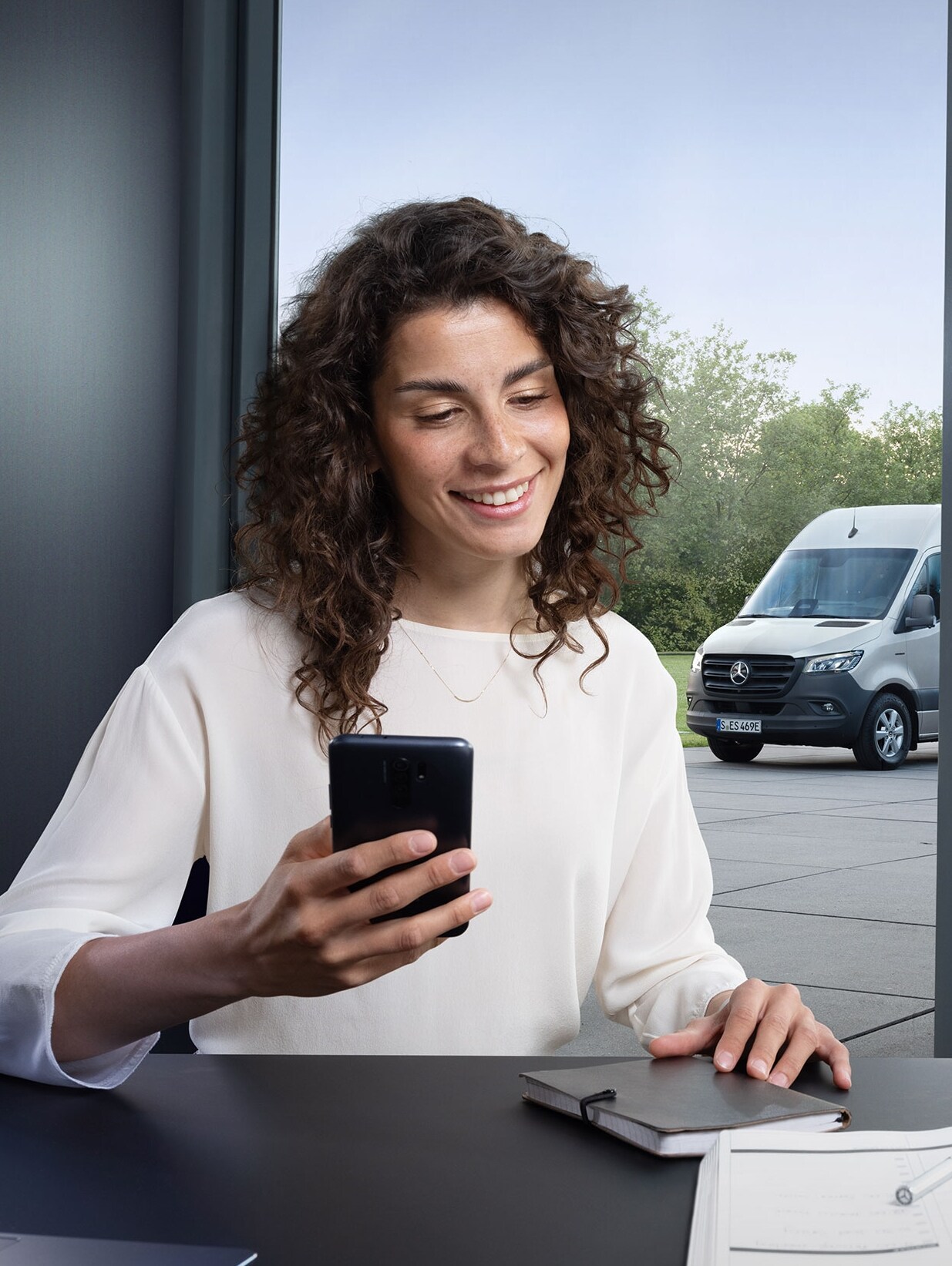 Vrouw die met een smartphone het wagenparkbeheer van meerdere Mercedes-Benz bestelwagens bedient bij het kantoorraam.