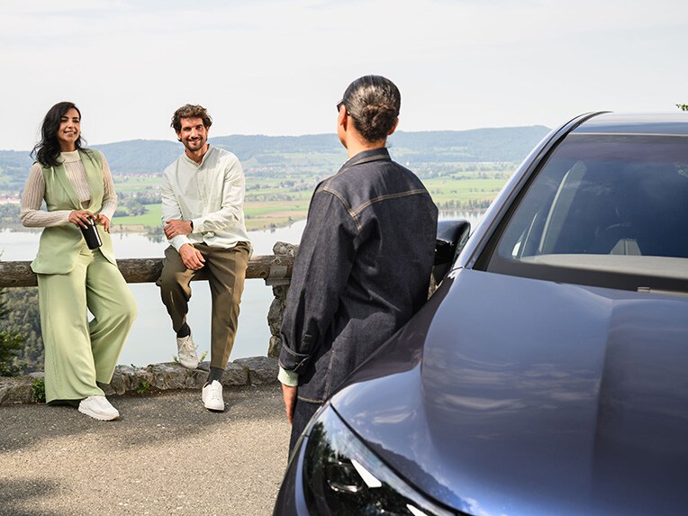 Een persoon leunt tegen een Mercedes-Benz voertuig en praat met twee andere personen die tegen een hek leunen.