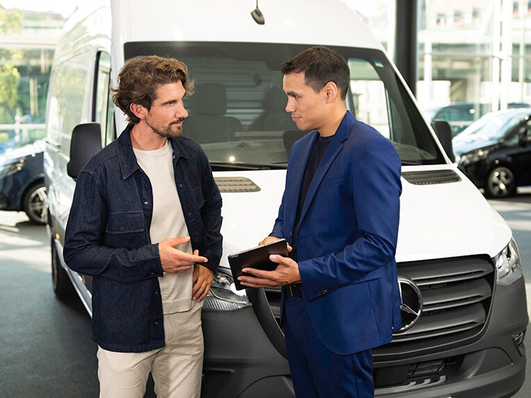 Twee personen zijn in gesprek en wijzen naar een mobiel apparaat terwijl ze voor een Mercedes-Benz bestelwagen staan.