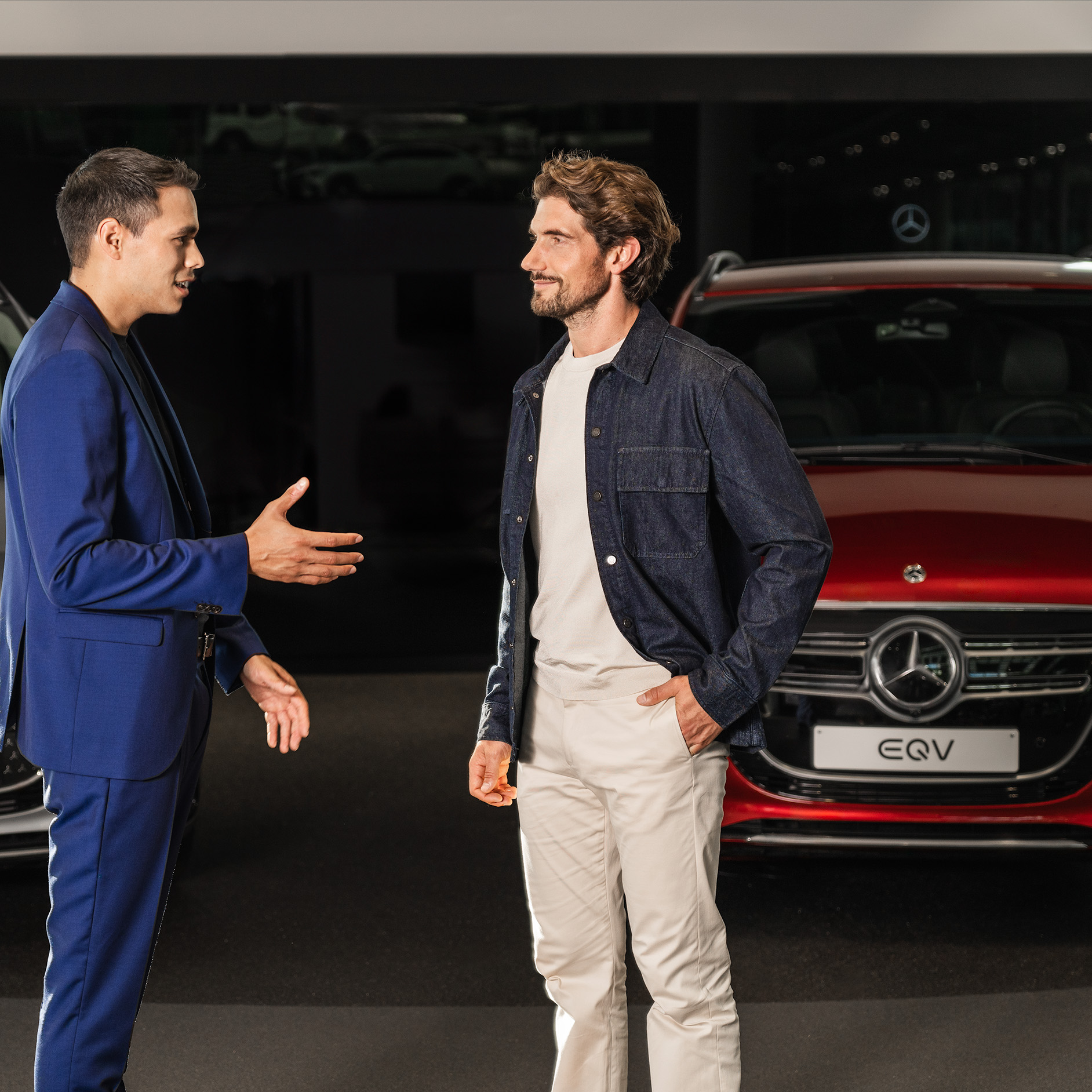 Twee mannen in gesprek in een Mercedes-Benz showroom met een rode EQV op de achtergrond. Twee mannen praten in een Mercedes-Benz showroom voor een rode EQV