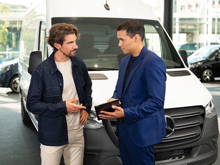 Twee personen zijn in gesprek en wijzen naar een mobiel apparaat terwijl ze voor een Mercedes-Benz bestelwagen staan.